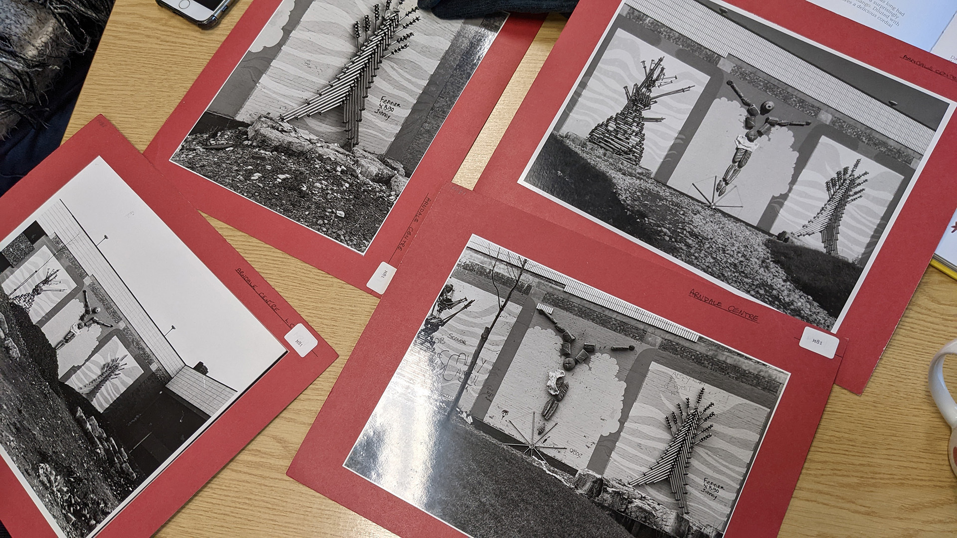 Photograph of several black and white photographs printed at A4, taped to a red background, and scattered o a table for people to look at. The photographs show a triptych wall art of what looks like a person next to two unusual sculptures.