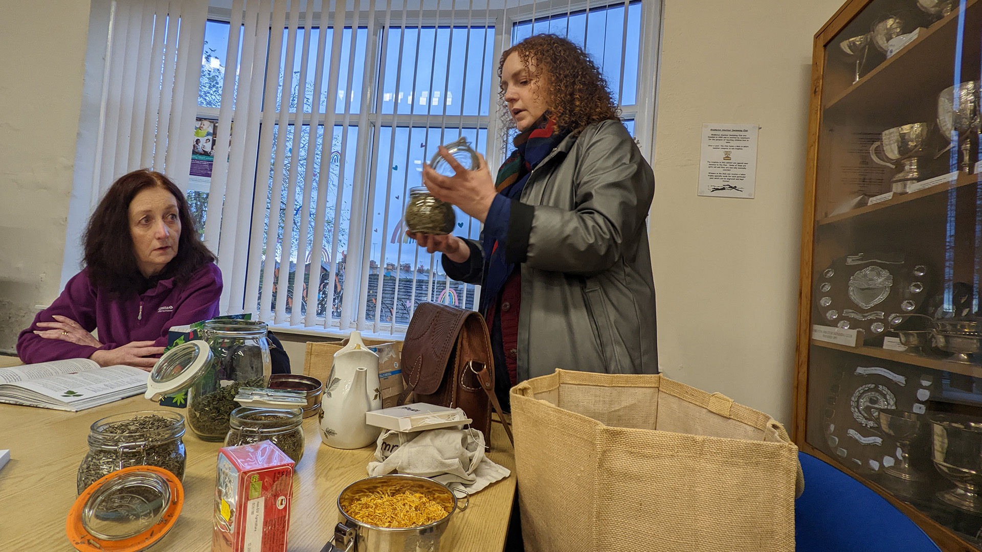 Photograph of the artist Alison Cooper leading a tea tasting session in a community building in Middleton. Alison holds up and talks about a particular type of tea, while another women at the table listens. The table is laden with other glass jars of tea and a ceramic tea pot.