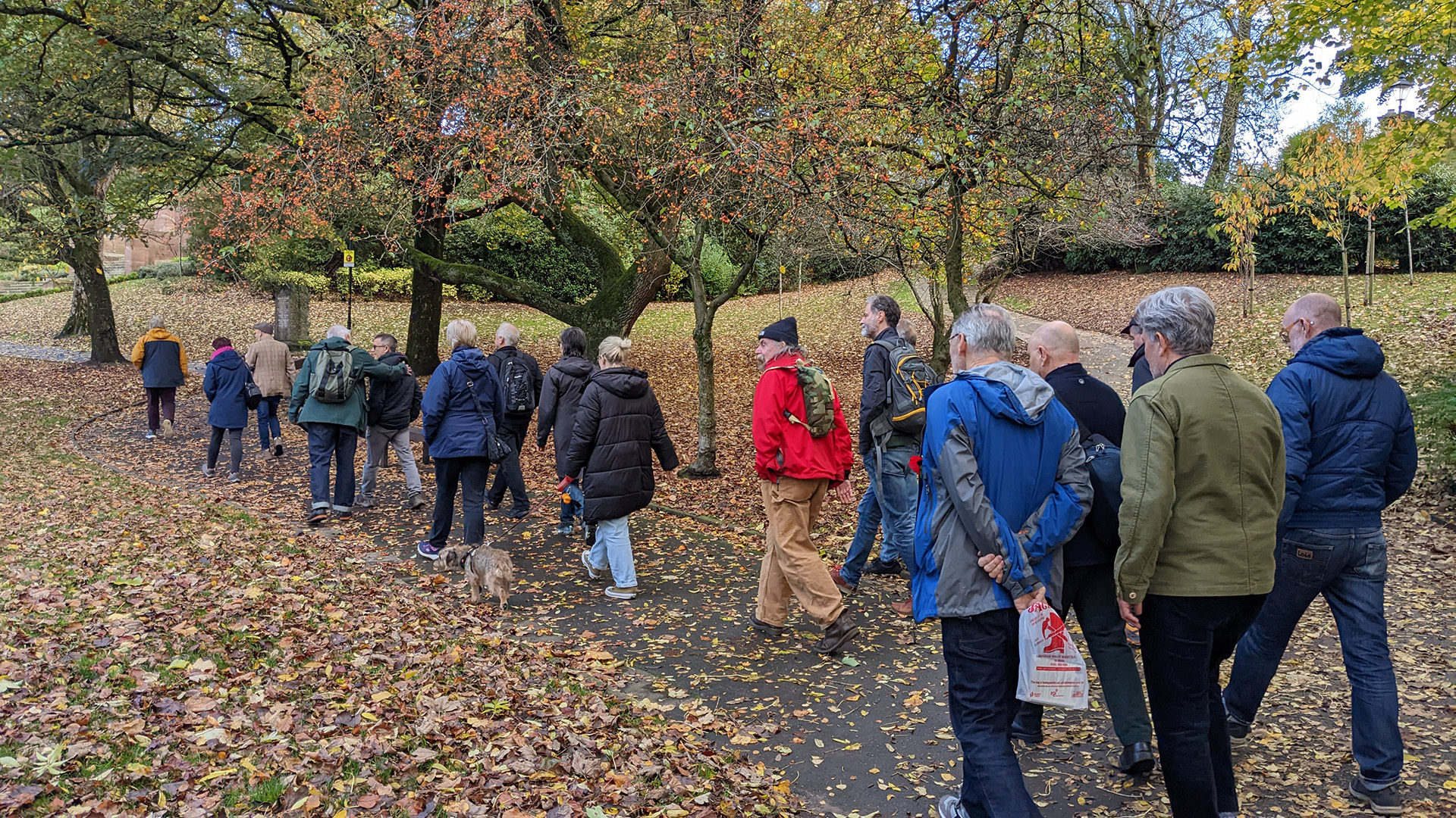 A photograph take during Stephen Marland's urban roam around Middleton. The image shows a group of people of around 15–20 people following Stephen along a park path. It is a bright day but the park is scattered with Autumn leaves.