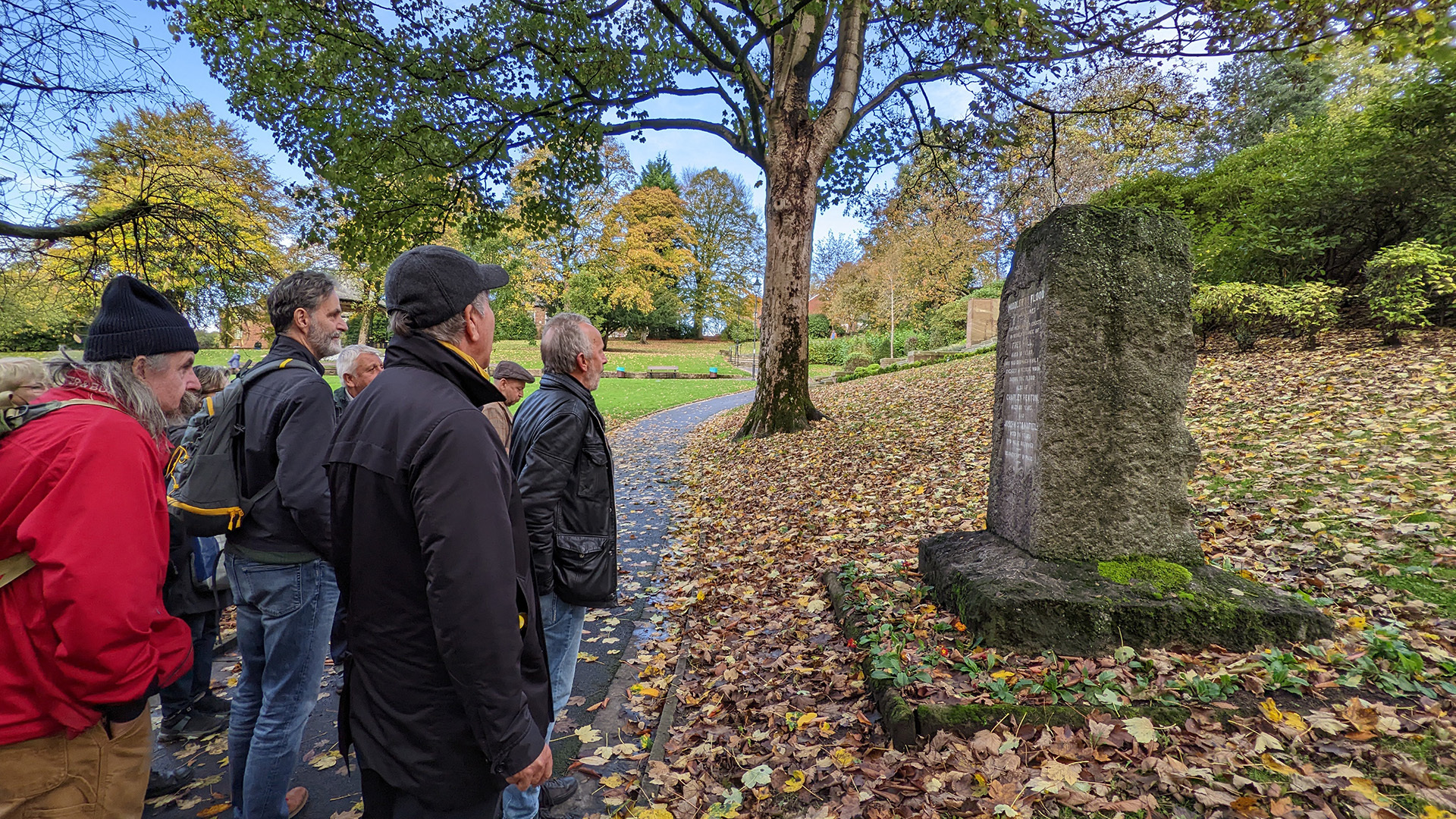 A photograph take during Stephen Marland's urban roam around Middleton. The image shows a group of people looking at a large commemorative stone, in a park scattered with autumn leaves. The stone has writing on it, but it is not legible in the photo.