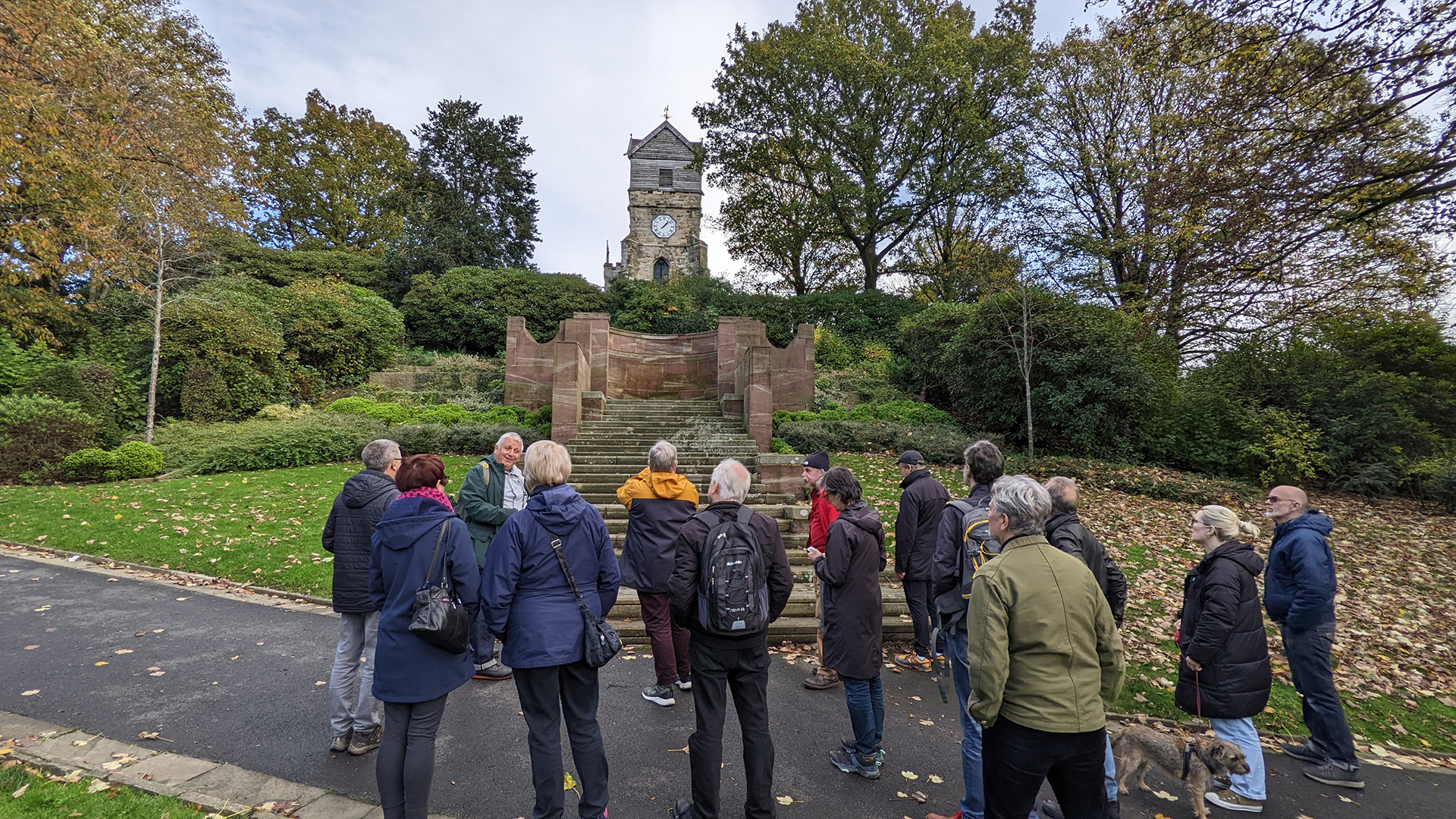 A photograph take during Stephen Marland's urban roam around Middleton. The image shows a group of around 15–20 people looking at a set of stone steps, in a park scattered with autumn leaves, as Stephen delivers his talk.