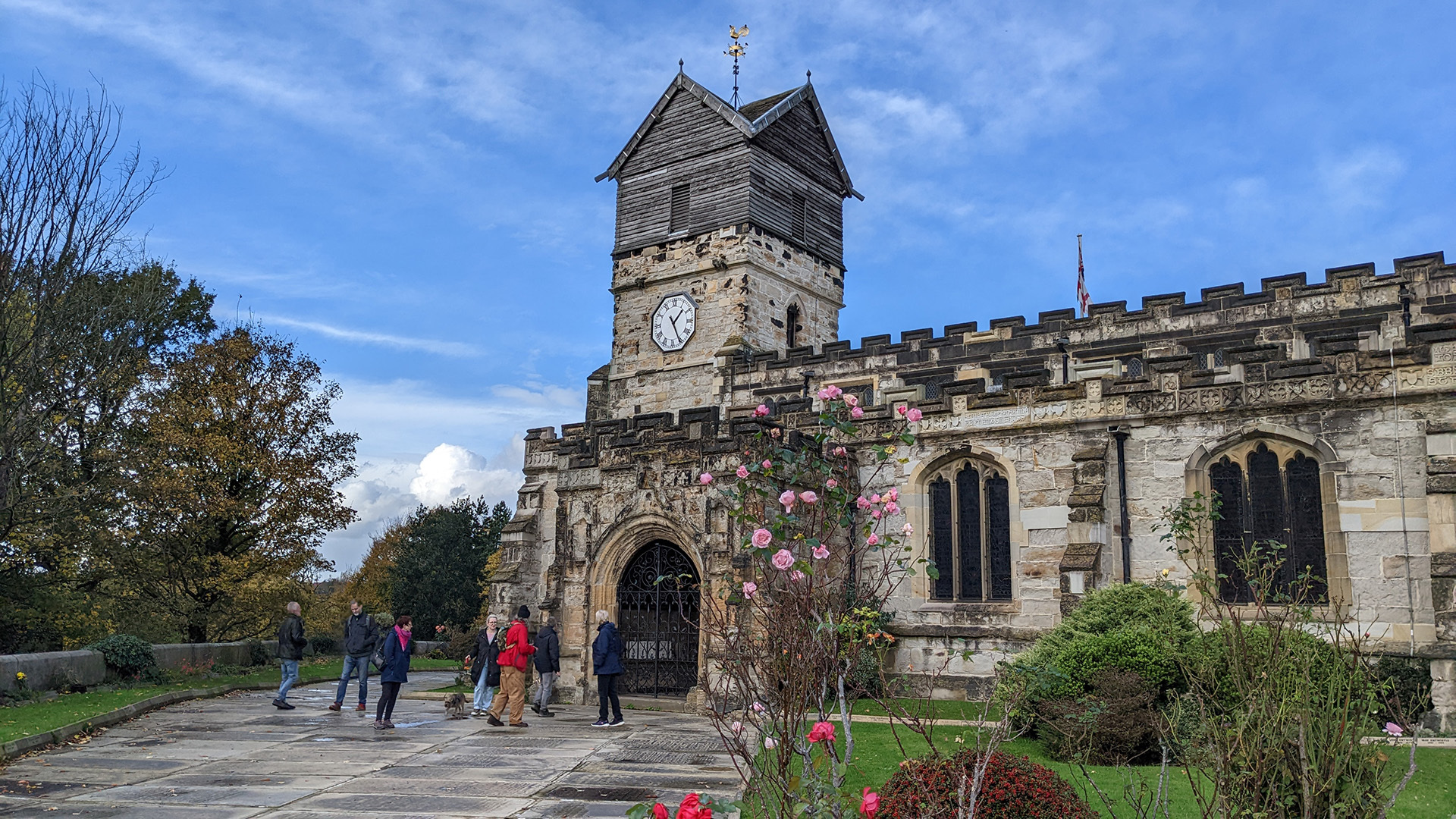 A photograph take during Stephen Marland's urban roam around Middleton. The image shows a group of people approaching an old church in the distance. The sky is clear and blue, and vibrant pink roses grow in the lush green grounds surrounding the church.