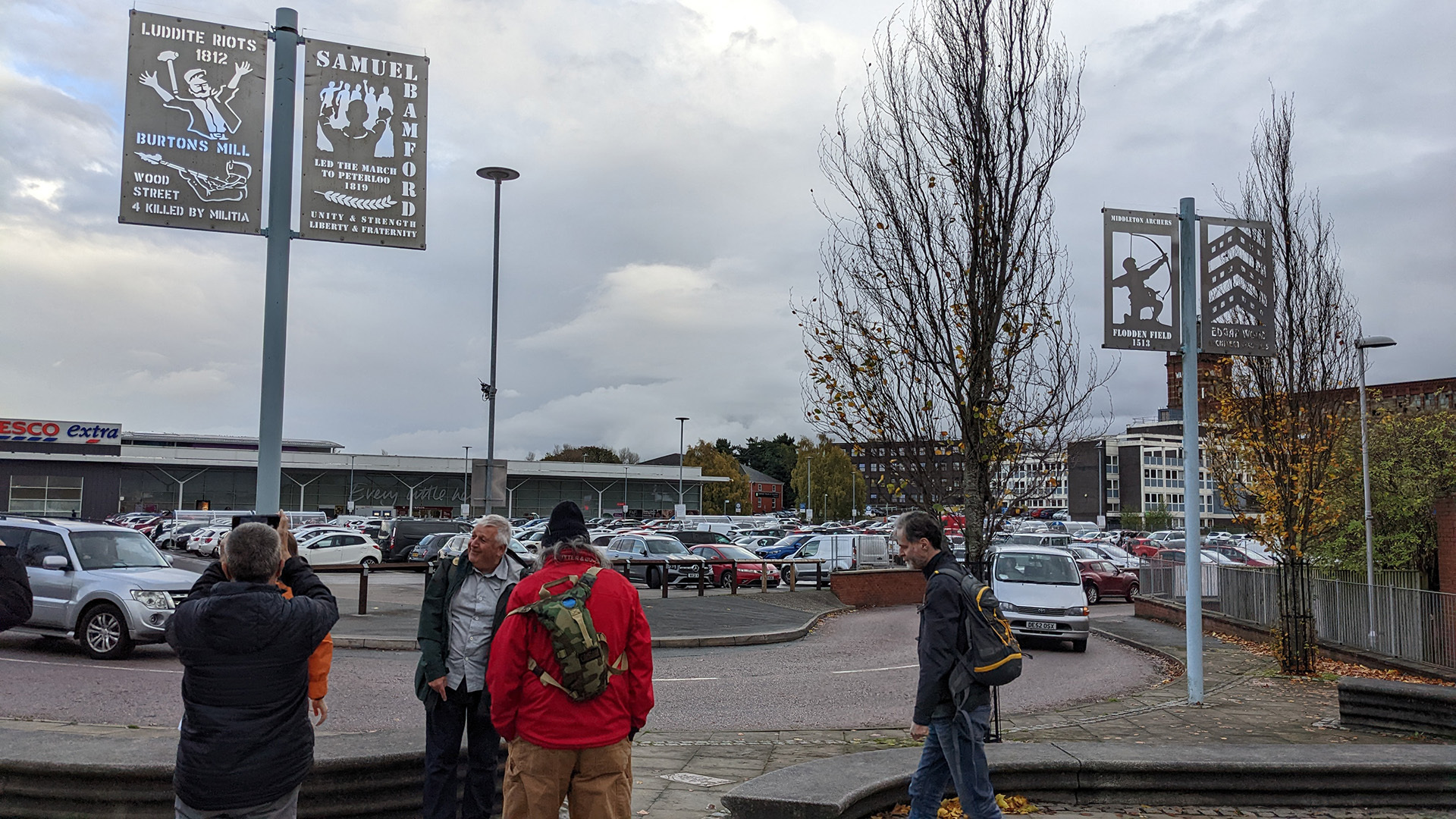 A photograph take during Stephen Marland's urban roam around Middleton. The image shows a group of people standing in a car park, looking up at artworks attached to the lampposts. The artworks are text and 2D illustrations cut out of rectangular metal sheets. Each one depicts a piece of local political history. The one closest to the camera depicts the Luddite Riots of 1812.