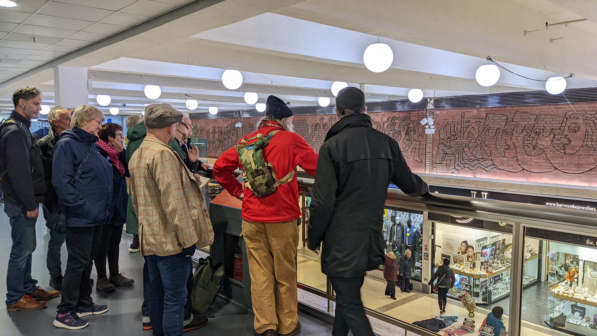 A photograph take during Stephen Marland's urban roam around Middleton. The image shows a group of people standing on the upper floor balcony of the local mall. They look over the balcony across to the wall on the far side, which displays decorative, red brick mosaic illustrations.